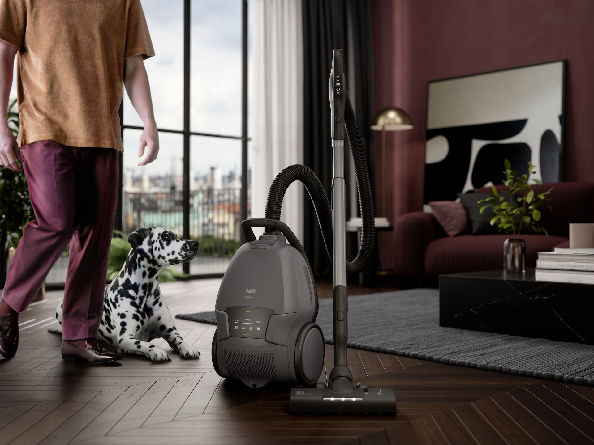 Een woonkamer met in beeld een vrouw die naar een AEG stofzuiger met zak loopt om te gaan stofzuigen. Er is ook een hond in beeld naast deze persoon.